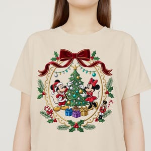May include: A beige t-shirt featuring a graphic of Mickey and Minnie Mouse decorating a Christmas tree inside a circular wreath. The design includes a red bow, holly leaves, candy canes, a nutcracker, and wrapped gifts in festive colors.