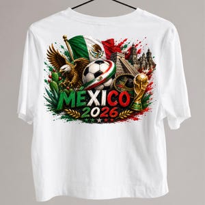 以下が含まれることがあります： メキシコを祝うカラフルなグラフィックデザインの白いTシャツ。デザインには、メキシコ国旗、サッカーボール、ワシ、そして緑と赤で「MEXICO 2026」の文字が含まれています。金のトロフィーとピラミッドも特徴です。
