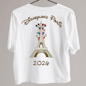 May include: White t-shirt with a gold design of the Eiffel Tower, Disney characters, and the text "Disneyland Paris 2026". The design includes Mickey and Minnie Mouse, Donald Duck, and other characters.