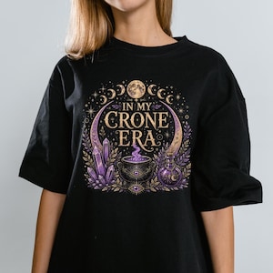 May include: A black t-shirt featuring a graphic print with the text IN MY CRONE ERA. The design includes a central cauldron with purple smoke, surrounded by purple crystals, a potion bottle, moon phases, and stars in gold and purple tones.