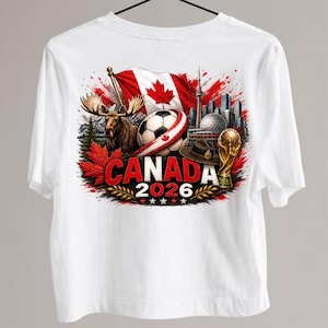 May include: White t-shirt featuring a graphic design with a Canadian theme. The design includes a Canadian flag, a soccer ball, a moose, and the text "CANADA 2026". The design also includes a trophy and a hat.