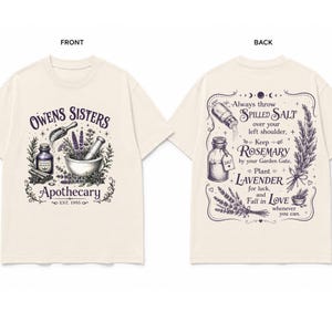 May include: A cream-colored t-shirt featuring a dark purple graphic. The front displays a mortar and pestle, herbs, and the text "Owens Sisters Apothecary Est. 1995." The back features text about salt, rosemary, and lavender with botanical illustrations.