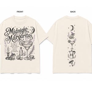 May include: A cream-colored t-shirt featuring a black ink illustration. The front displays a margarita glass, a frog in a witch hat, a candle, and the text Midnight Margaritas. The back shows a vertical design with a moon, frog, glass, and mushroom.