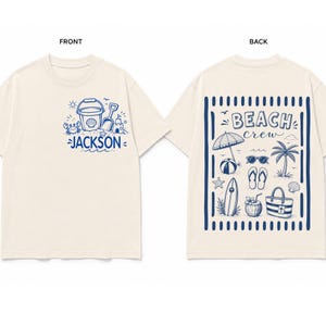 Puede incluir: Una camiseta de manga corta color crema con ilustraciones de líneas azules. El frente muestra un cubo, una pala, un cangrejo y el nombre JACKSON. La parte posterior presenta una escena de playa con sombrilla, tabla de surf y el texto BEACH crew.