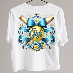 May include: White t-shirt featuring a softball design. The design includes a yellow softball with the name "Gigi" in script, crossed baseball bats, and blue ribbons. The word "Softball" is above the design.
