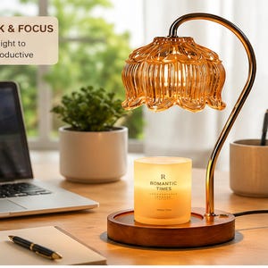 May include: A candle warmer lamp with a curved gold-tone metal arm and a translucent amber glass shade shaped like a flower. The base is circular wood, holding a white candle jar labeled "ROMANTIC TIMES." The lamp emits a warm, soft glow.