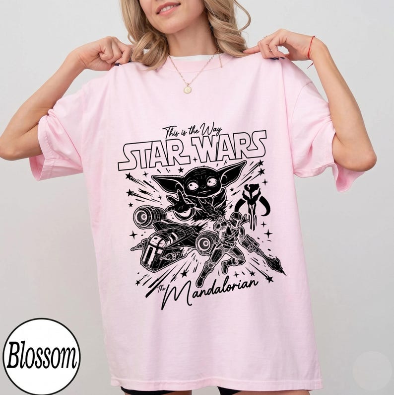 May include: A light pink t-shirt featuring a black graphic print of Grogu, a Mandalorian warrior, a starship, and the text "This is the Way STAR WARS The Mandalorian". The design includes star accents and line art detailing across the centre chest area.