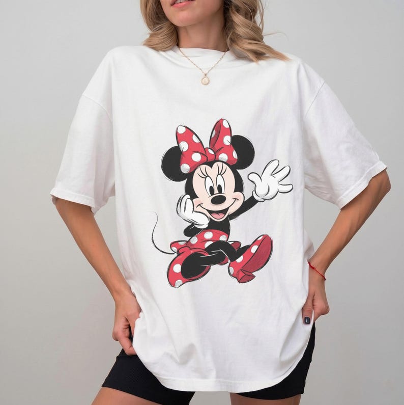 May include: A white short-sleeved t-shirt featuring a large graphic print of Minnie Mouse on the front. The character is depicted in her signature red and white polka dot dress and bow, with black ears and white gloves, centered on the chest.
