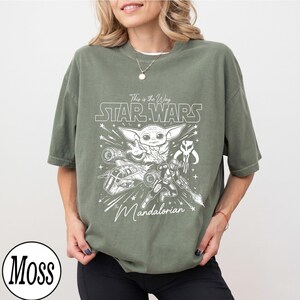 May include: An olive green short-sleeve t-shirt featuring a white graphic print of Grogu, a Mandalorian warrior, a starship, and the text "This is the Way STAR WARS The Mandalorian". The shirt has a crew neckline and a relaxed, loose-fitting silhouette.