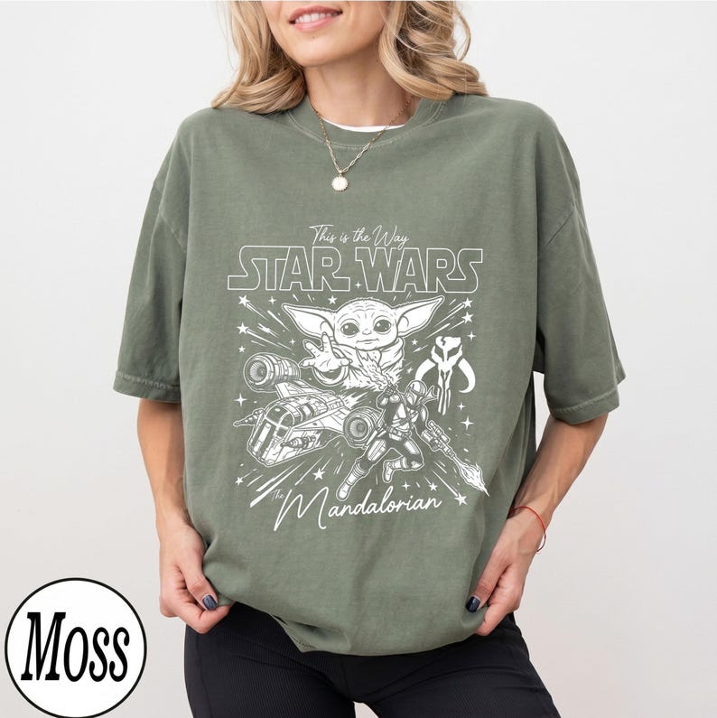 May include: An olive green short-sleeve t-shirt featuring a white graphic print of Grogu, a Mandalorian warrior, a starship, and the text "This is the Way STAR WARS The Mandalorian". The shirt has a crew neckline and a relaxed, loose-fitting silhouette.