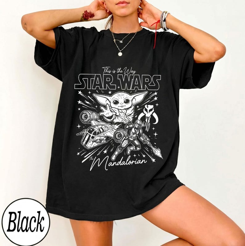 May include: A black short-sleeved t-shirt featuring a white graphic print of Grogu, a Mandalorian warrior, a starship, and the text This is the Way, Star Wars, and The Mandalorian. The design includes starburst accents on a dark fabric background.