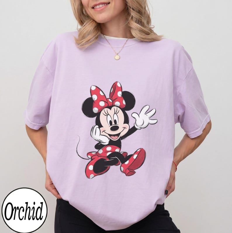 May include: A light purple short-sleeve t-shirt featuring a graphic print of Minnie Mouse wearing a red and white polka dot bow and matching dress. The character is depicted in a cheerful pose with white gloves and red shoes on a solid background.