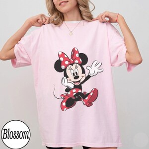 May include: A light pink short-sleeved t-shirt featuring a graphic print of Minnie Mouse wearing a red and white polka dot bow and matching dress. The character is depicted in a classic pose with one hand raised. A circular logo reads Blossom.