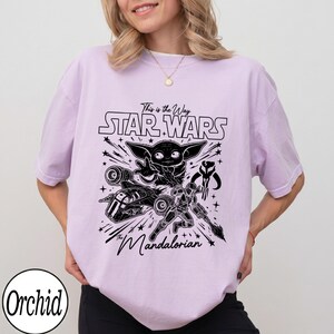 May include: A light purple t-shirt featuring a black graphic print with the text "This is the Way", "STAR WARS", and "The Mandalorian". The design includes illustrations of Grogu, a Mandalorian character, a spaceship, and a helmet symbol.