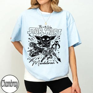 May include: A light blue t-shirt featuring a black graphic print with the text This is the Way, STAR WARS, and The Mandalorian. The design includes illustrations of Grogu, a Mandalorian character, a spaceship, and a helmet icon surrounded by stars.