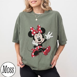May include: An olive green short-sleeve t-shirt featuring a graphic print of Minnie Mouse. The character is depicted with her signature red and white polka dot bow and matching dress, waving with one hand. The shirt has a crew neckline.