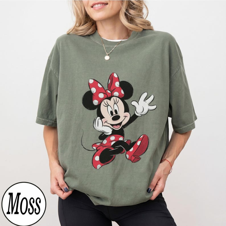 May include: An olive green short-sleeve t-shirt featuring a graphic print of Minnie Mouse. The character is depicted with her signature red and white polka dot bow and matching dress, waving with one hand. The shirt has a crew neckline.