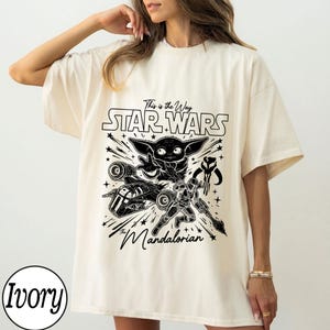 May include: An ivory short-sleeve t-shirt featuring a black graphic print of Grogu, a Mandalorian warrior, a starship, and the text "This is the Way", "STAR WARS", and "The Mandalorian". The design includes various star shapes and lines.