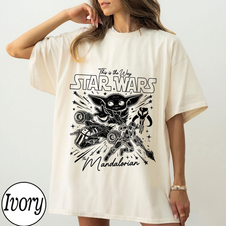 May include: An ivory short-sleeve t-shirt featuring a black graphic print of Grogu, a Mandalorian warrior, a starship, and the text "This is the Way", "STAR WARS", and "The Mandalorian". The design includes various star shapes and lines.