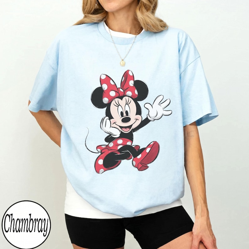 May include: A light blue t-shirt featuring a graphic of Minnie Mouse wearing a red and white polka dot bow and dress. The character is depicted in a classic pose with one hand raised. The shirt has short sleeves and a crew neckline.