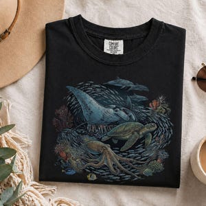 Op de afbeelding: Een zwart t-shirt met een gedetailleerde grafische print van een mantarog, een zeeschildpad en een octopus die tussen koraal en een school vissen zwemmen. Het shirt heeft een Comfort Colors merklabel in de kraag en een ronde halslijn.