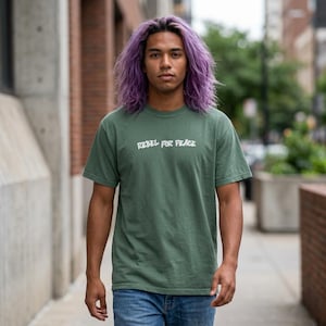 Op de afbeelding: Een olijfgroen T-shirt met korte mouwen en de witte tekst REBEL FOR PEACE op de borst gedrukt. Het kledingstuk heeft een ronde hals en een standaard pasvorm, gemaakt van een effen gekleurde stof met een eenvoudig en strak ontwerp.