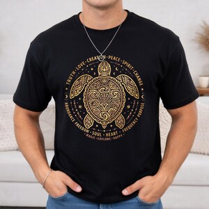 Puede incluir: Una camiseta negra de manga corta con un gráfico de color dorado de una tortuga marina centrado en el pecho. La tortuga está rodeada por un borde circular que contiene las palabras Truth, Love, Creation, Peace, Spirit, Change, Purpose, Frequency, Happy, Explore, Magic, Heart, Soul, Freedom y Abundance.