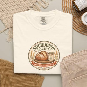 Op de afbeelding: Een crèmekleurig t-shirt met een cirkelvormige afbeelding met de tekst SOURDOUGH SOCIETY, KNEAD REALITY, BAKE BREAD. Het ontwerp bevat een brood op een houten plank en een glazen pot met het label Live Starter, omringd door korenaren.