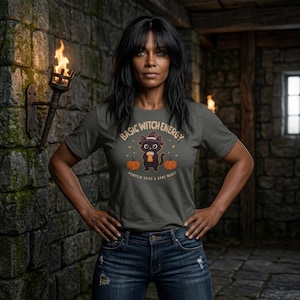 Op de afbeelding: Een donkergrijs t-shirt met korte mouwen en een afbeelding van een zwarte kat met een heksenhoed die een koffiebeker vasthoudt, geflankeerd door twee oranje pompoenen. De tekst luidt "BASIC WITCH ENERGY" en "PUMPKIN SPICE & DARK MAGIC".