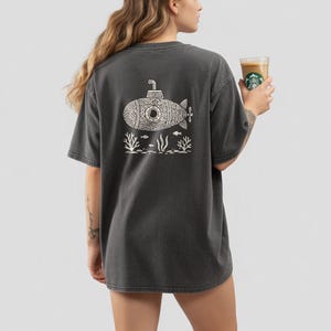 Puede incluir: Una camiseta de manga corta en color gris carbón con un gráfico de líneas blancas en la espalda. El diseño muestra un submarino rodeado de algas y peces pequeños. La prenda tiene cuello redondo y un corte holgado y relajado.