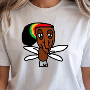 Op de afbeelding: Een wit t-shirt met een grafische illustratie van een bruin figuur met een lange neus en hangende ogen. Het figuur draagt een zwarte, rode, gele en groene muts en heeft vier witte vleugels aan de zijkanten, gecentreerd op de borst.