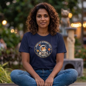 Op de afbeelding: Een marineblauw t-shirt met een cirkelvormige afbeelding van een oranje cyperse kat in een witte astronautenhelm en ruimtepak. De tekst luidt COSMIC CAT ASTRONOMY GUILD en EXPLORING THE PURR-VERSE, omringd door kleine sterren en planeten.