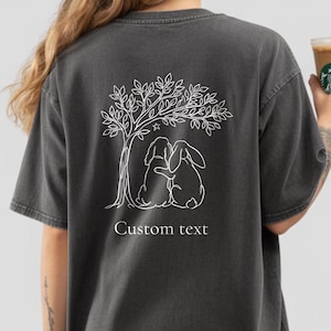 Op de afbeelding: Een antracietgrijs t-shirt met korte mouwen en een witte lijntekening op de achterkant. Het ontwerp toont een boom met twee dieren eronder, waarvan er één zijn arm om de ander slaat, en een kleine ster. De tekst Custom text staat eronder.