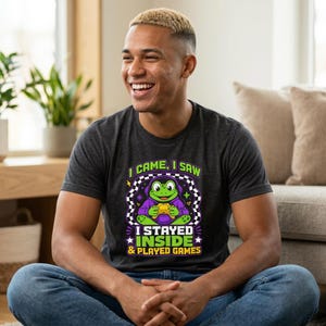 Op de afbeelding: Een donkergrijs t-shirt met een afbeelding van een groene kikker met een paarse koptelefoon die een gele gamecontroller vasthoudt. De tekst luidt I CAME, I SAW I STAYED INSIDE & PLAYED GAMES in witte, gele en groene letters.