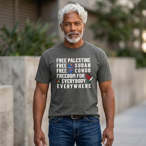 Op de afbeelding: Een antracietgrijs t-shirt met korte mouwen en witte tekst: FREE PALESTINE, FREE SUDAN, FREE CONGO, FREEDOM FOR EVERYBODY EVERYWHERE. Het ontwerp bevat een klein blauw, geel en rood vlagpictogram en een kleine vogelafbeelding.