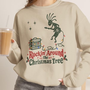 Op de afbeelding: Een beige sweater met ronde hals en een donkergroene afbeelding van een Kokopelli-figuur die op een elektrische gitaar speelt. Het ontwerp bevat een gitaarversterker, kleurrijke kerstverlichting en de tekst Rockin' Around The Christmas Tree.
