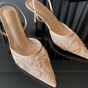 Puede incluir: Un par de zapatos de tacón beige con punta. Los zapatos presentan intrincados bordados florales dorados y una delicada correa en el tobillo. Los tacones miden aproximadamente 5 cm de alto.