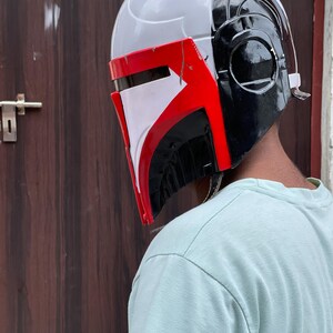 May include: A Mandalorian helmet with a white, red, and black color scheme. The helmet has a sleek design with a visor and ear pieces. The red accents are on the visor and the side of the helmet. The helmet is a cosplay item.