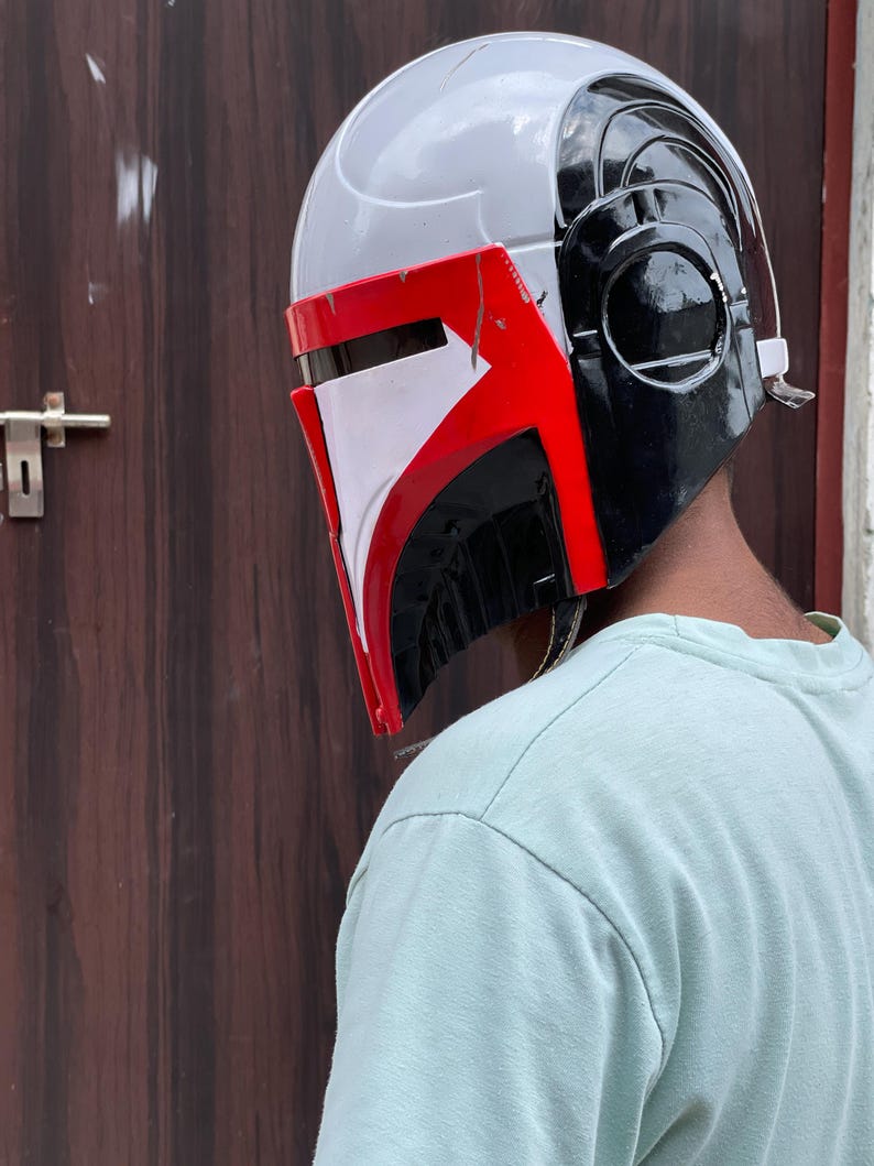 May include: A Mandalorian helmet with a white, red, and black color scheme. The helmet has a sleek design with a visor and ear pieces. The red accents are on the visor and the side of the helmet. The helmet is a cosplay item.