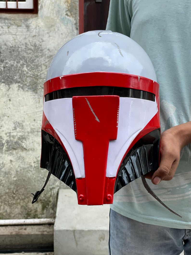 May include: A gray, red, white, and black helmet with a distinctive design. The helmet features a rounded top, a red visor area, and white and black accents. The helmet is held by a person.