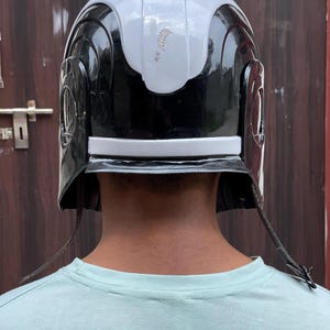 May include: A black and white helmet with a silver stripe. The helmet has a rounded top and side details. The helmet has two leather straps hanging down on each side.