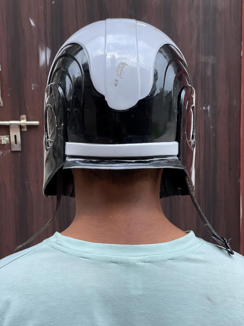 May include: A black and white helmet with a silver stripe. The helmet has a rounded top and side details. The helmet has two leather straps hanging down on each side.