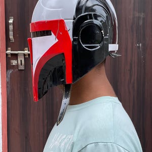 May include: A Mandalorian helmet in a side profile view. The helmet is primarily white and black, with red accents on the visor. It has a sleek, futuristic design with a leather strap.
