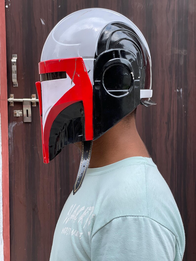 May include: A Mandalorian helmet in a side profile view. The helmet is primarily white and black, with red accents on the visor. It has a sleek, futuristic design with a leather strap.