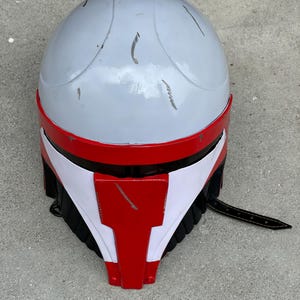 May include: A gray and red helmet with white accents and a black strap. The helmet has a rounded top and a red visor. Scratches are visible on the surface. The helmet appears to be a costume or prop.