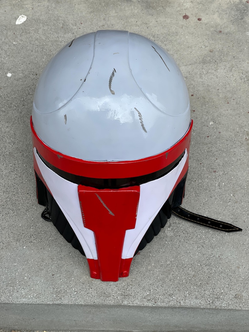May include: A gray and red helmet with white accents and a black strap. The helmet has a rounded top and a red visor. Scratches are visible on the surface. The helmet appears to be a costume or prop.