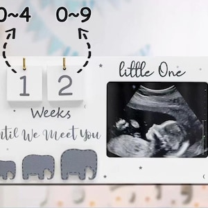 May include: A white rectangular wooden frame featuring a photo slot for an ultrasound, two hanging blocks displaying the number 12, and the text "little One" and "Weeks Until We Meet You". Three grey elephant silhouettes are at the bottom.
