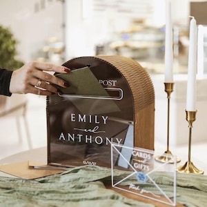 Op de afbeelding: Een houten kaartenbox met een gebogen bovenkant en een donker acryl voorpaneel met de witte tekst "POST", "EMILY and ANTHONY" en "12/12/27". Een klein doorzichtig acrylbordje met "CARDS and GIFTS" staat voor de box.