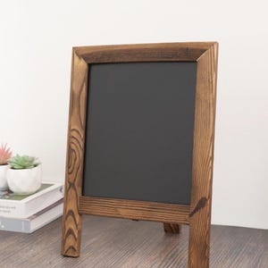 May include: A rectangular chalkboard featuring a dark, smooth writing surface framed by natural, stained wood with visible grain patterns. The frame is constructed with mitered corners and stands upright on two sturdy, integrated wooden legs.