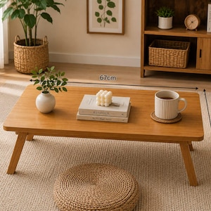 May include: A rectangular wooden coffee table with four angled legs, measuring 67 cm in length and 34 cm in height. The table surface holds a white vase with green leaves, two stacked books, a white bubble candle, and a white ribbed mug on a coaster.